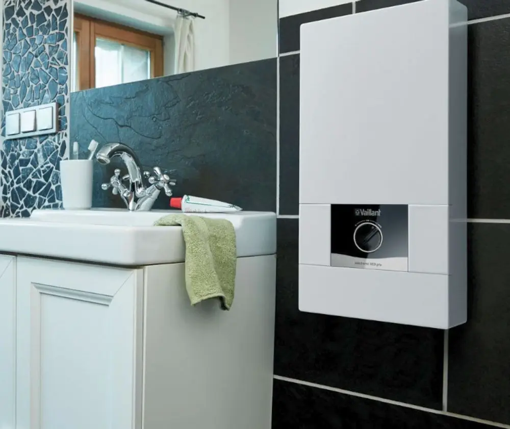 Vaillant Elektro-Durchlauferhitzer an Wand montiert, neben Waschbecken mit einem Handtuch, im Bad mit dunklem Wanddesign und fensterseitiger Beleuchtung.
